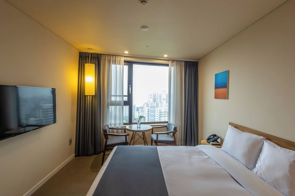 A hotel room with a bed, a TV, a desk with two chairs, a window with curtains, and a painting on the wall.