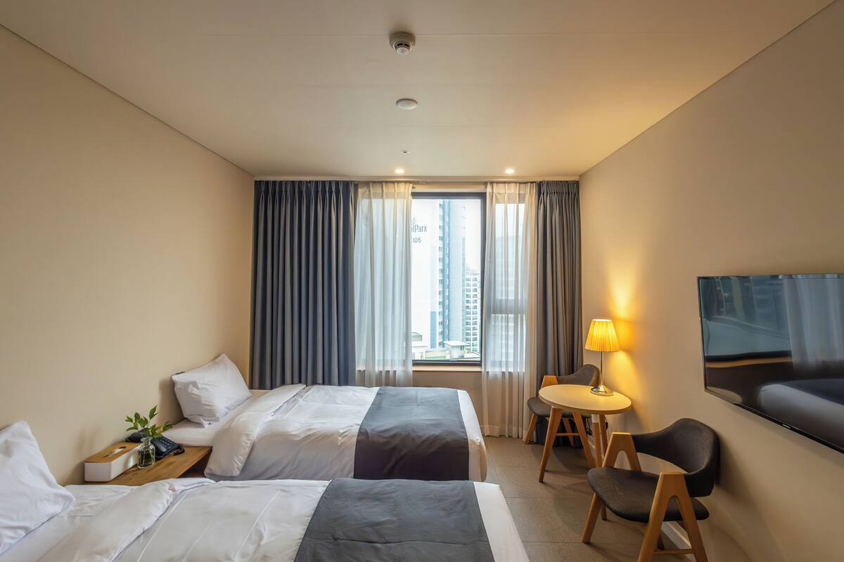 A hotel room with two beds, a desk with a lamp, a TV, and a view of a cityscape through the window.