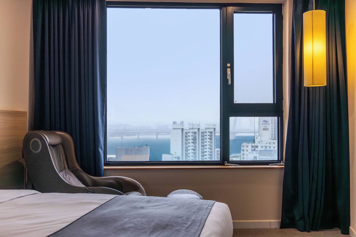 A hotel room with a bed, a massage chair, a large window with curtains, and a view of a cityscape.