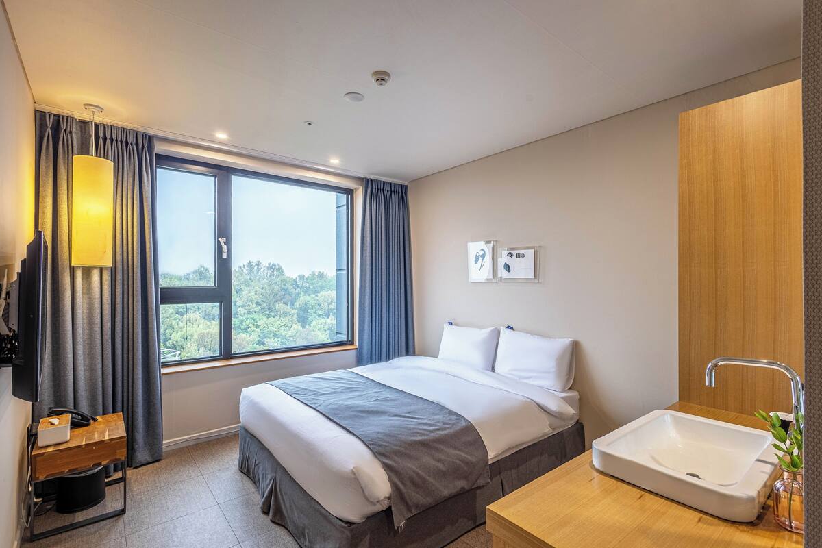 A modern hotel room with a large bed, a bedside table, a built-in sink, and a view of the outdoors.
