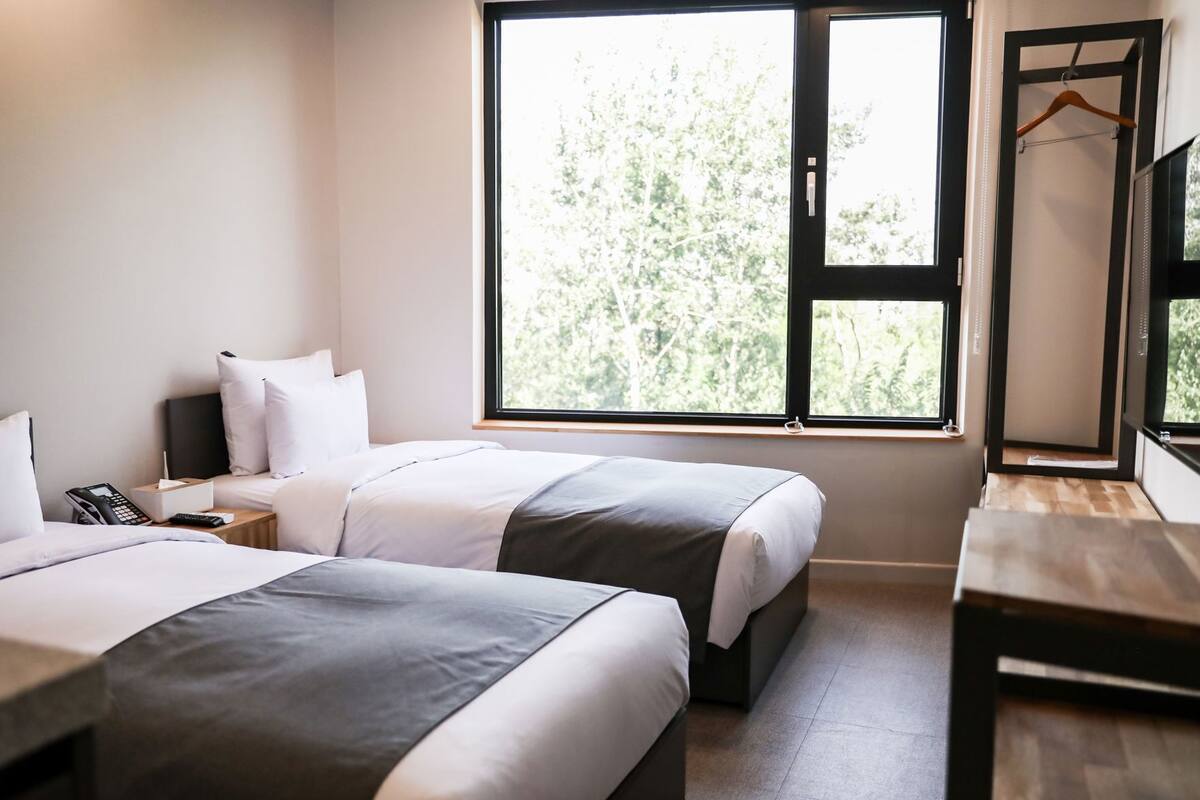 A hotel room with two beds, a bedside table with a phone, a bedside lamp, and a window with a view of trees.