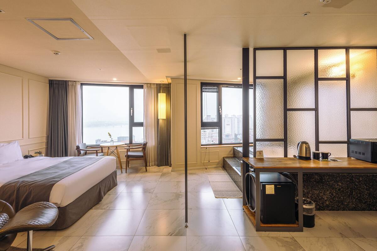 A hotel room with a bed, desk, kitchenette, and large windows offering a city view.