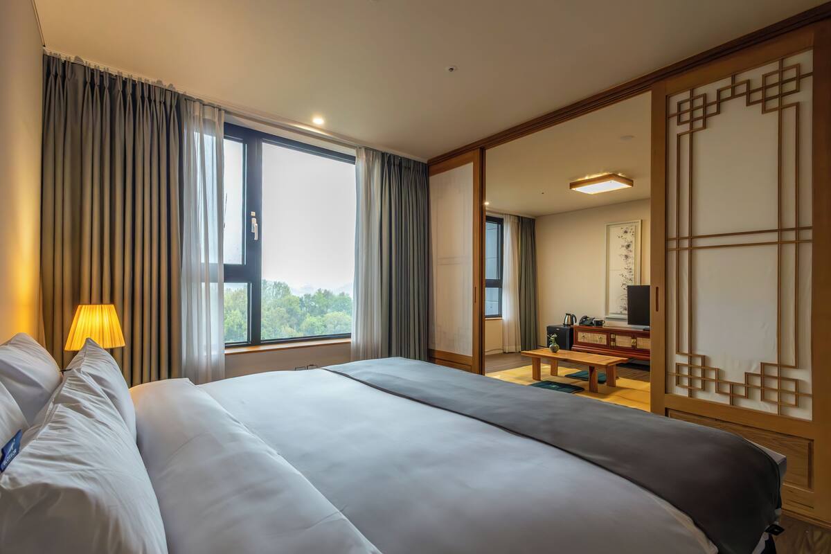 A hotel room with a large bed, a desk, a chair, a lamp, and a view of the outdoors.
