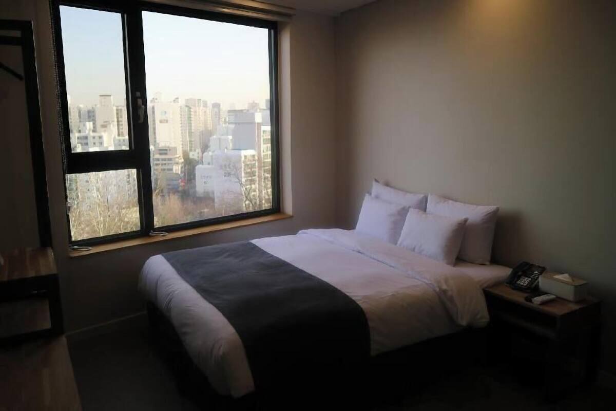 A hotel room with a bed, a nightstand, a window with a city view, and a door.
