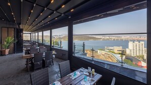 An indoor dining area with tables set for two, large windows offering a view of a bridge and cityscape. - JK Blossom Hotel (Seoul)