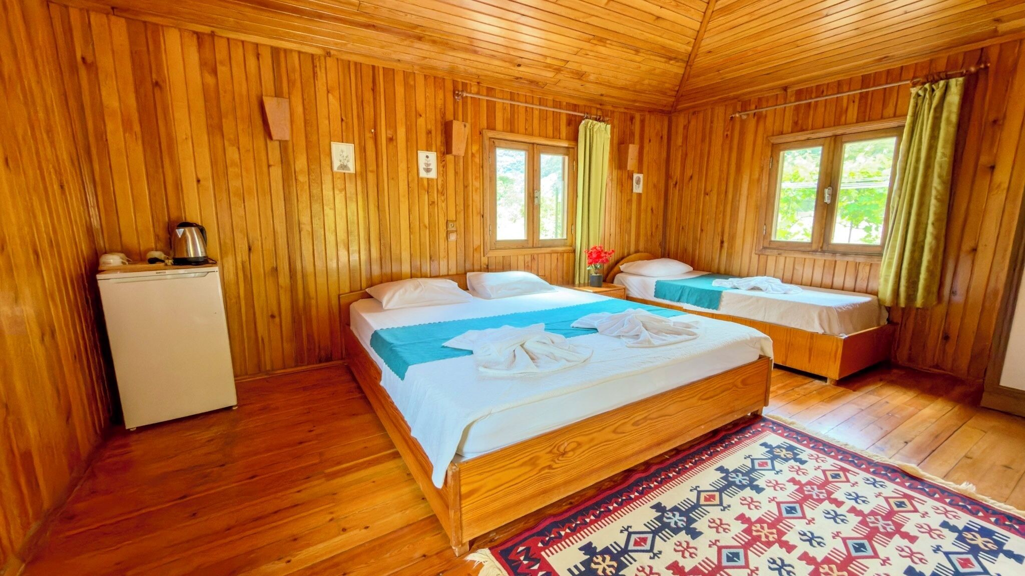 A wooden cabin with two beds, a small kitchenette, and a window with curtains.