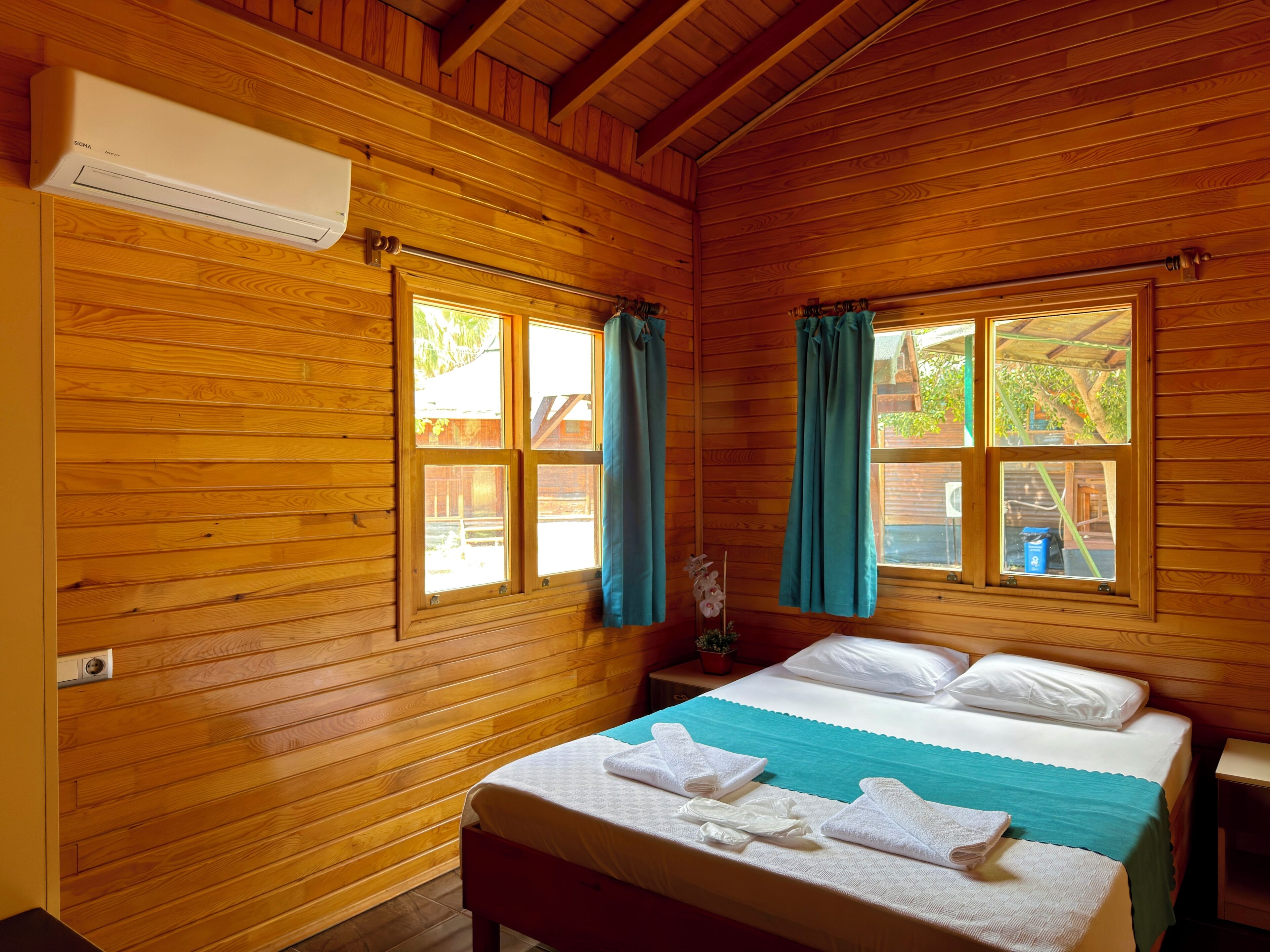 Una habitación de cabaña de madera con una cama, dos ventanas con cortinas y un aire acondicionado de pared.