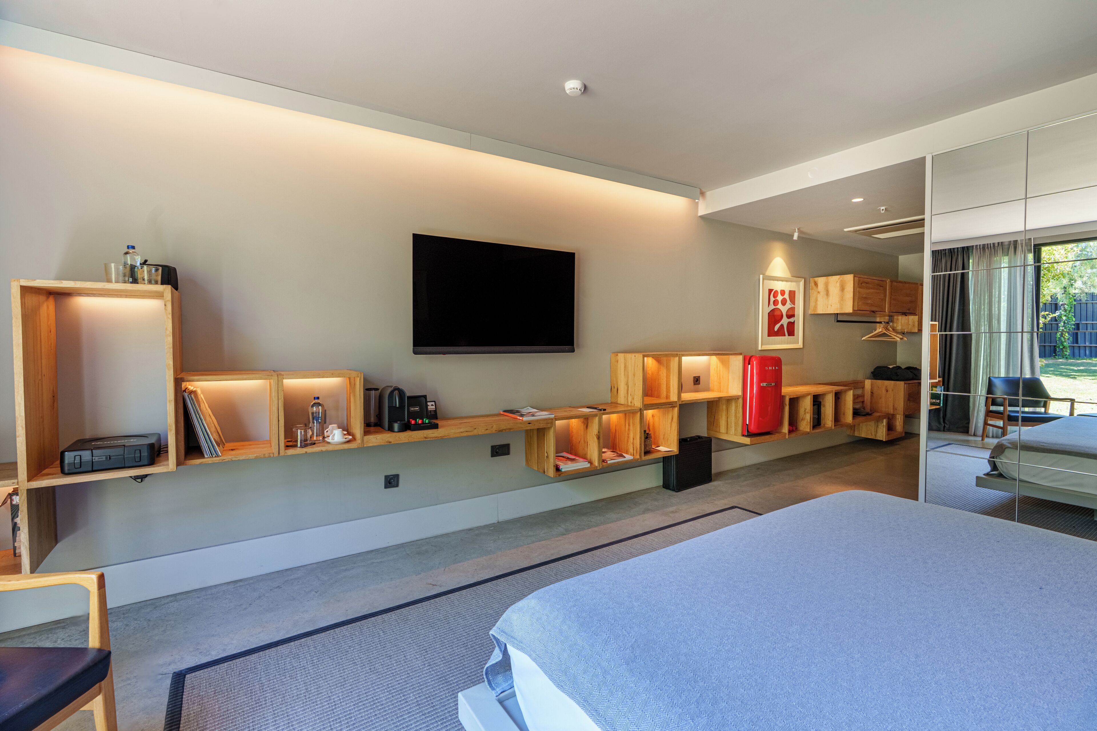 A modern hotel room with a large bed, built-in shelves with lighting, a flat-screen TV, and a view of the outdoors.