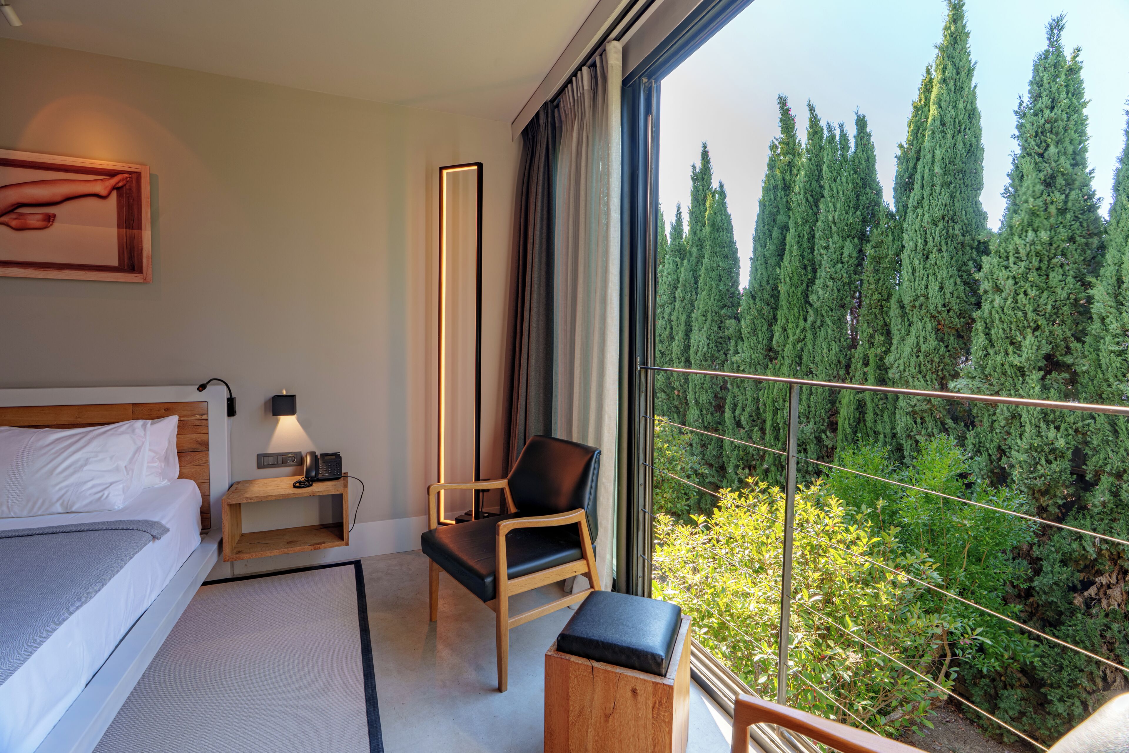 A hotel room with a bed, a chair, a nightstand, a bedside table, and a large window offering a view of trees.