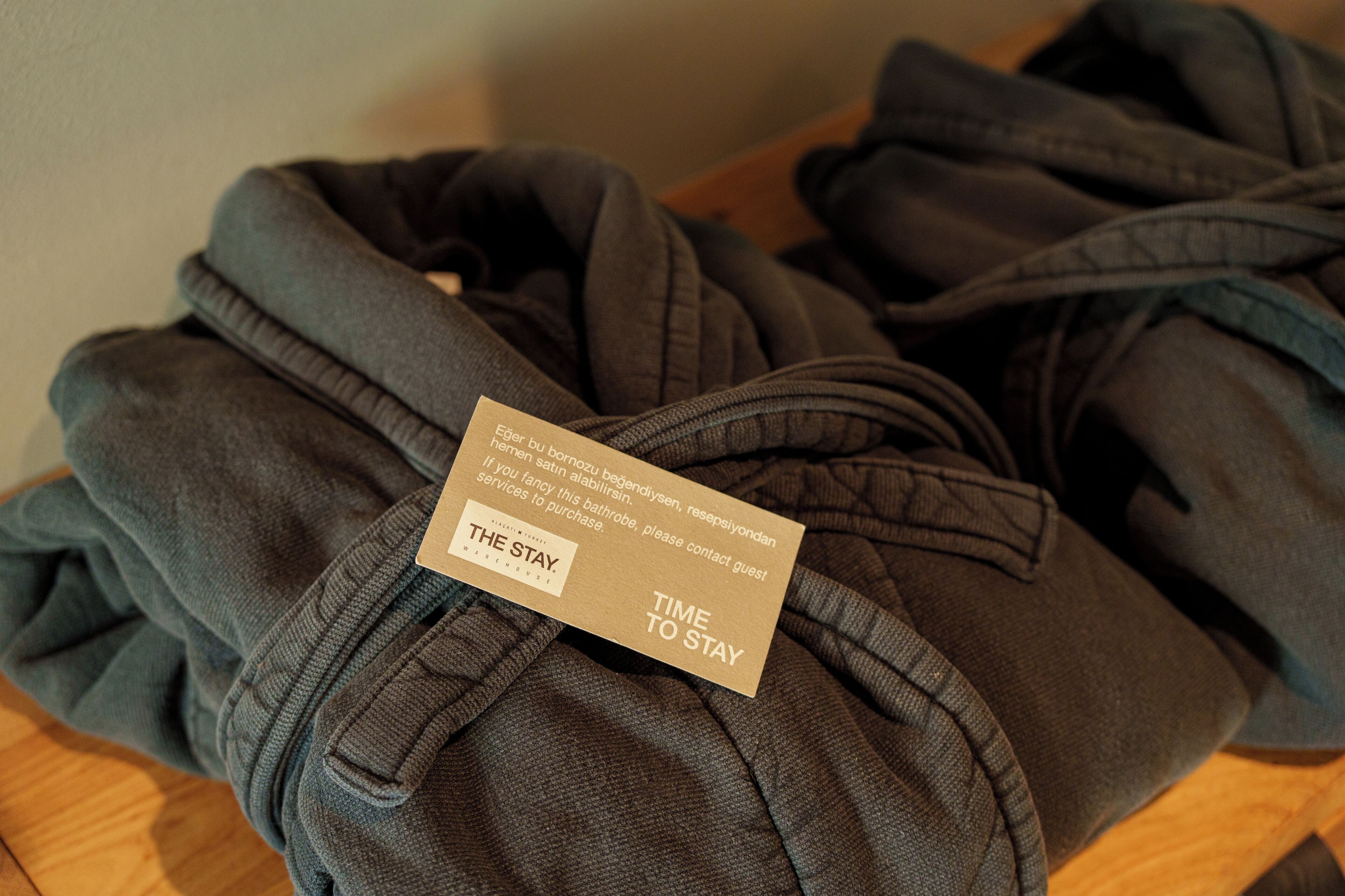 Folded dark-colored robes on a wooden surface with a tag that reads 'TIME TO STAY' and 'THE STAY.'