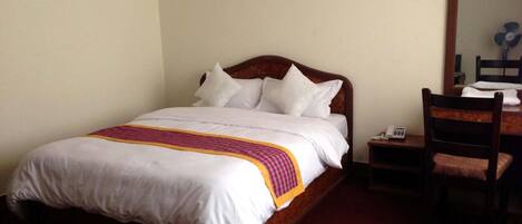 A neatly made bed with white linens and a pink and yellow striped blanket. There is a wooden nightstand with a telephone beside the bed. A wooden chair with a cushion is positioned next to the nightstand.