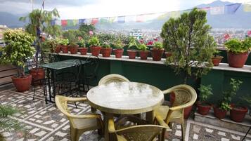 A terrace with a round table and chairs, surrounded by potted plants and a view of a cityscape.