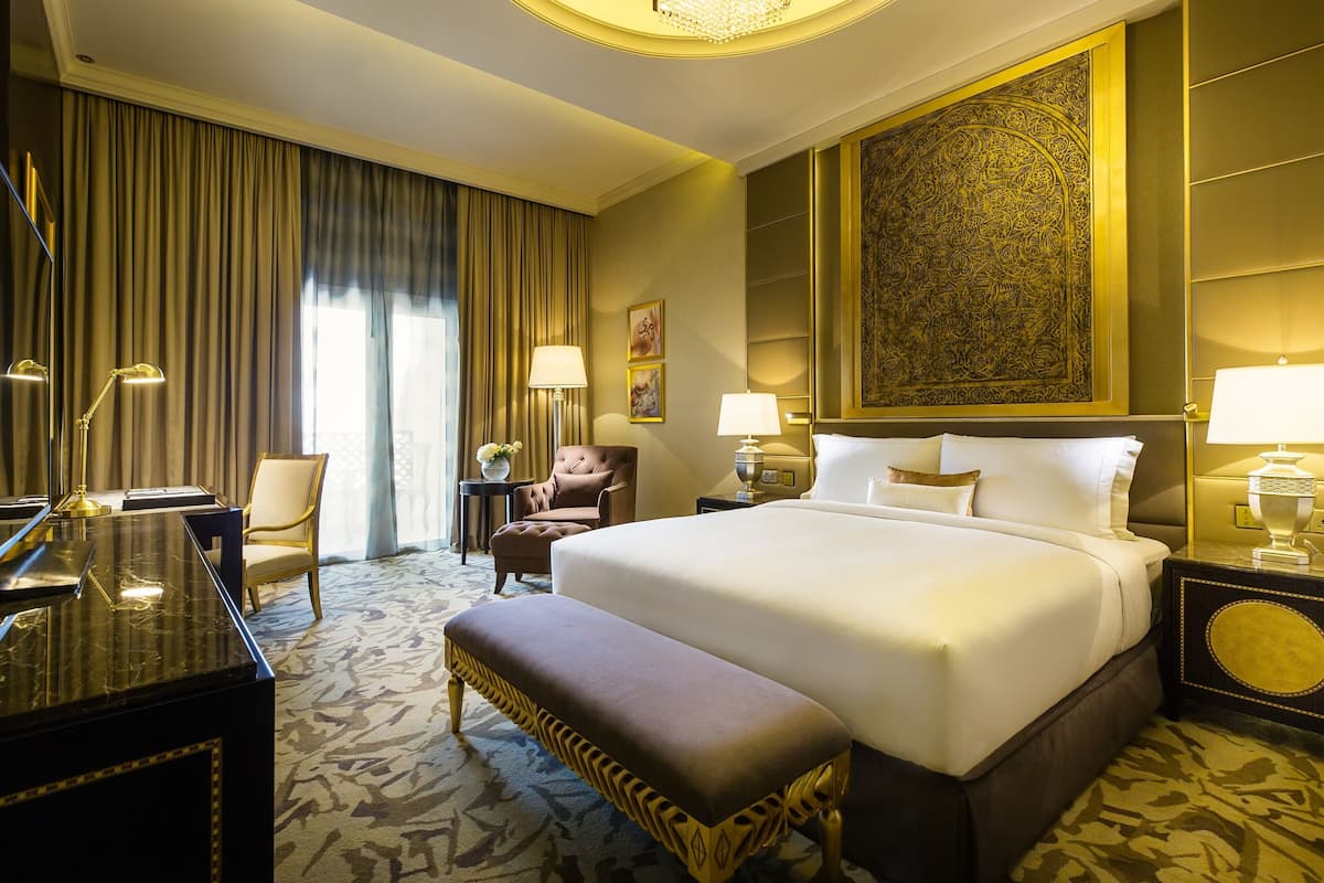 A hotel room with a large bed, a desk, a chair, a bench, and a large decorative wall piece.