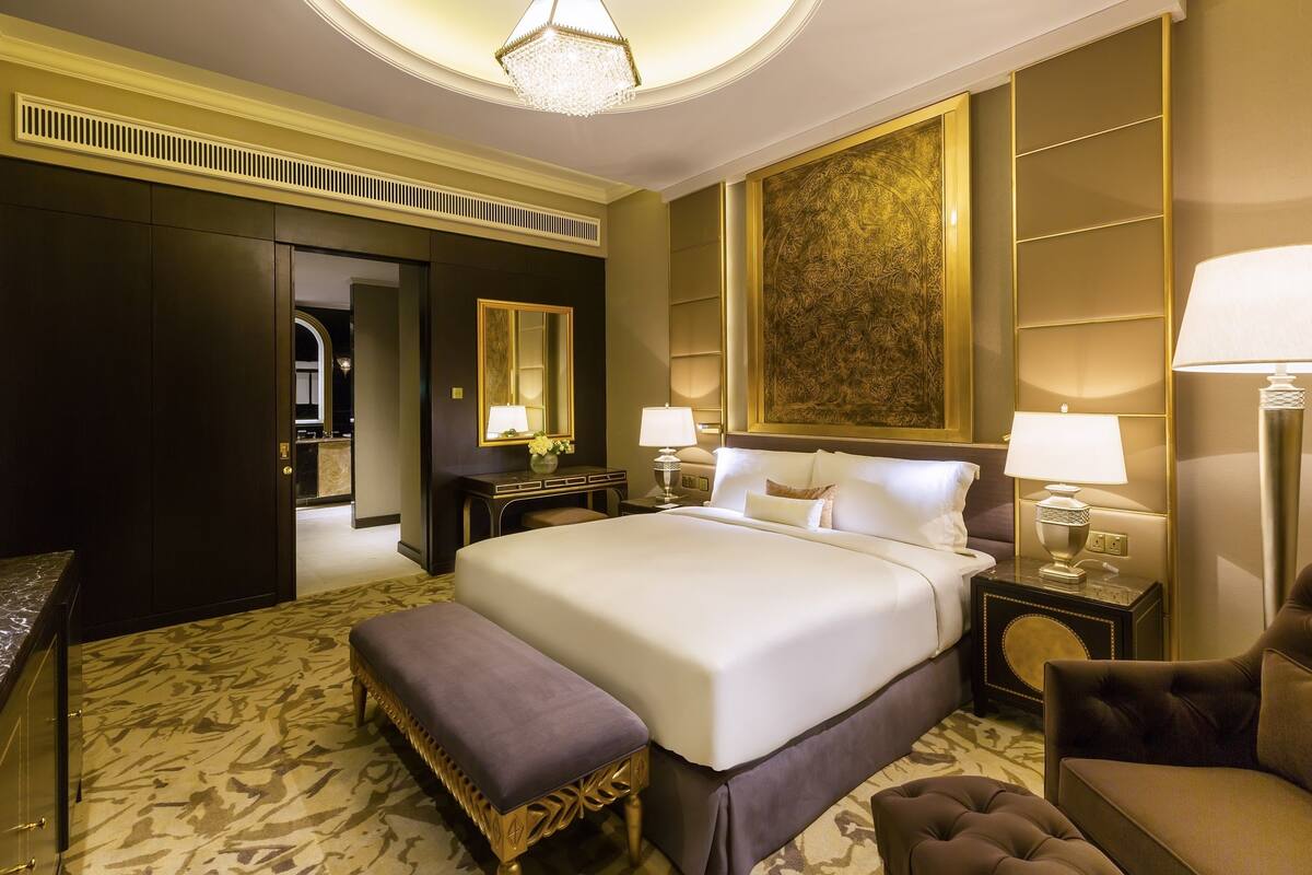 A spacious hotel room with a large bed, a sitting area with a bench, a desk, and a mirror.