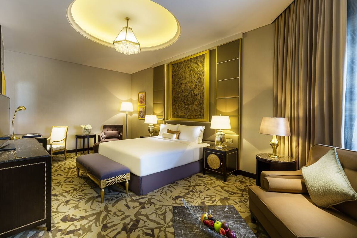 A hotel room with a large bed, two bedside tables with lamps, a sofa, a desk, and a patterned carpet.