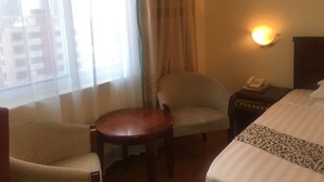 Une chambre d’hôtel avec un lit, une chaise, une petite table, une table de chevet avec un téléphone et une fenêtre avec des rideaux.