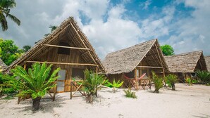 A row of thatched-roof huts with a sandy beach and palm trees in the foreground. - New Jambo Bungalows - Hostel (Paje)