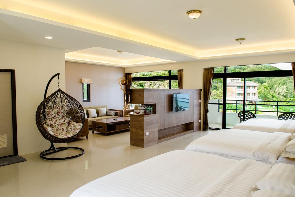 Une chambre d’hôtel dotée d’un grand lit, d’un coin salon avec un canapé et un fauteuil suspendu, d’un téléviseur fixé au mur et d’un balcon offrant une vue sur la verdure environnante.