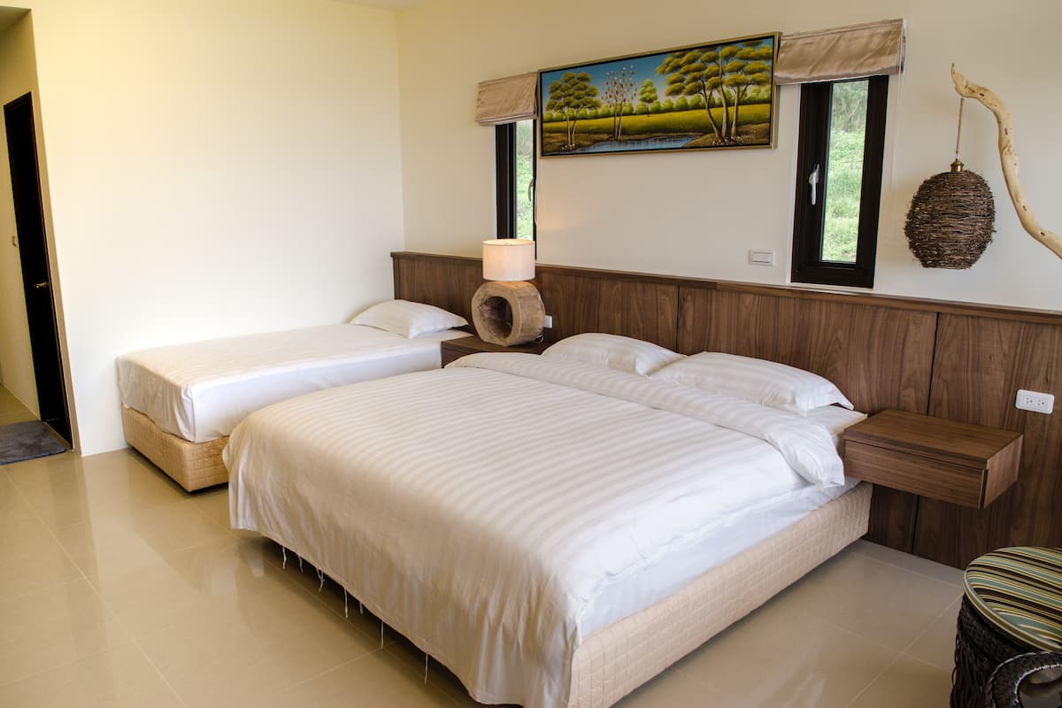 Une chambre avec deux lits, une tête de lit en bois, un tableau représentant des arbres, un luminaire suspendu et une fenêtre donnant sur un paysage verdoyant.