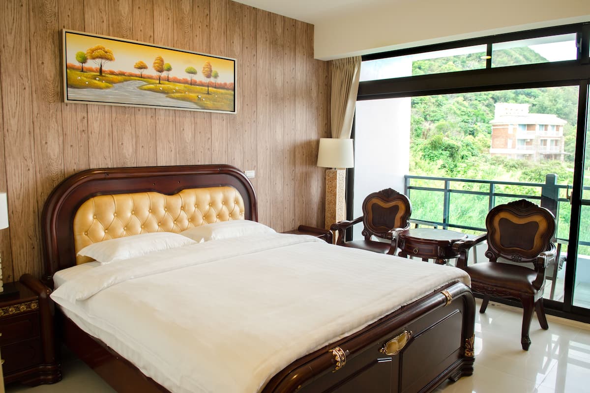 Une chambre d’hôtel avec un grand lit, une tête de lit en bois, un tableau représentant un paysage, un balcon donnant sur un espace verdoyant et une table avec des chaises.