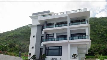 A multi-story building with balconies, a sign reading "HOLA", and a lush green hillside in the background.