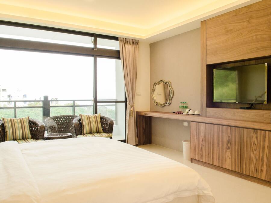A hotel room with a large bed, a TV mounted on a wooden cabinet, a balcony with wicker chairs, and a view of the city.