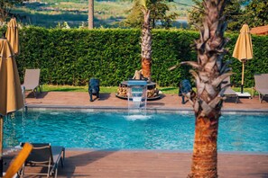 Ein Schwimmbecken mit Liegestühlen und Sonnenschirmen, umgeben von Grün und mit Blick auf die Landschaft.