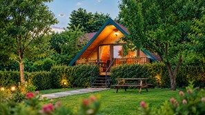 Une cabane en bois avec un escalier menant à une terrasse, entourée d’une végétation luxuriante et d’une pelouse bien entretenue.
