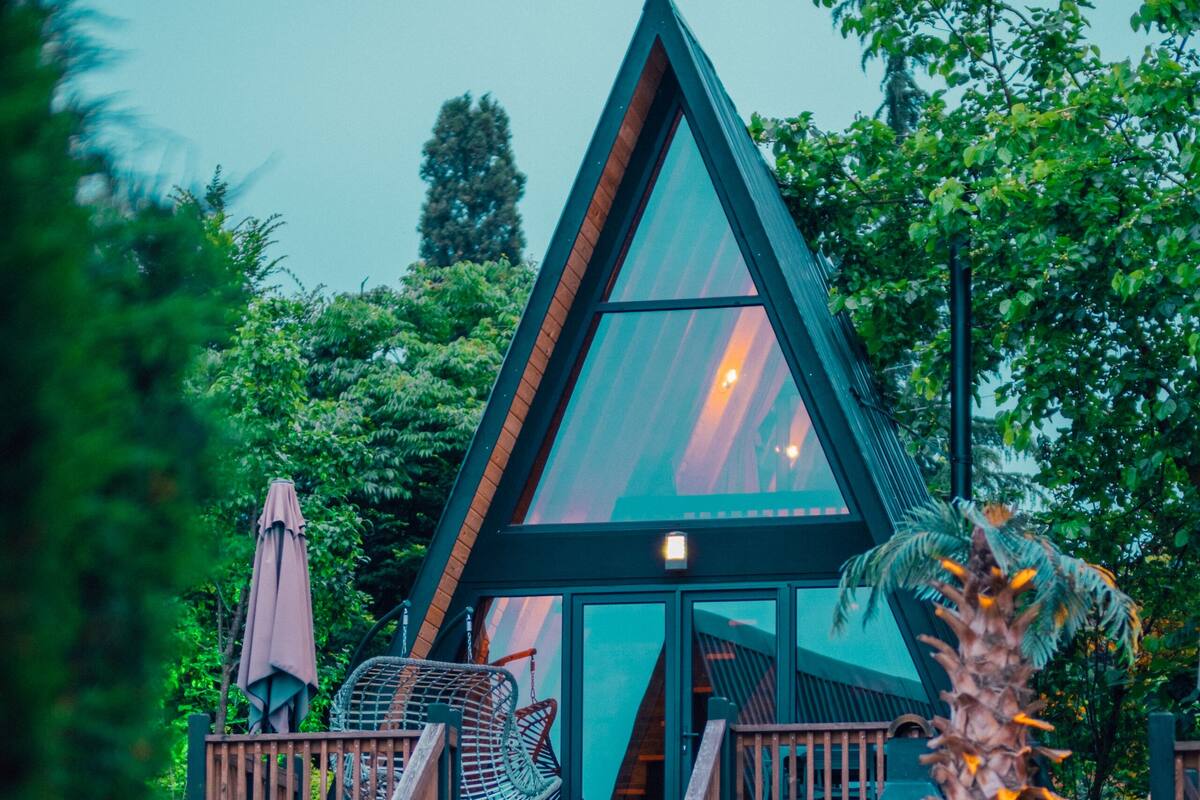 Un bâtiment moderne de forme triangulaire, doté d’une terrasse en bois et de grandes fenêtres en verre, et entouré d’une végétation luxuriante.