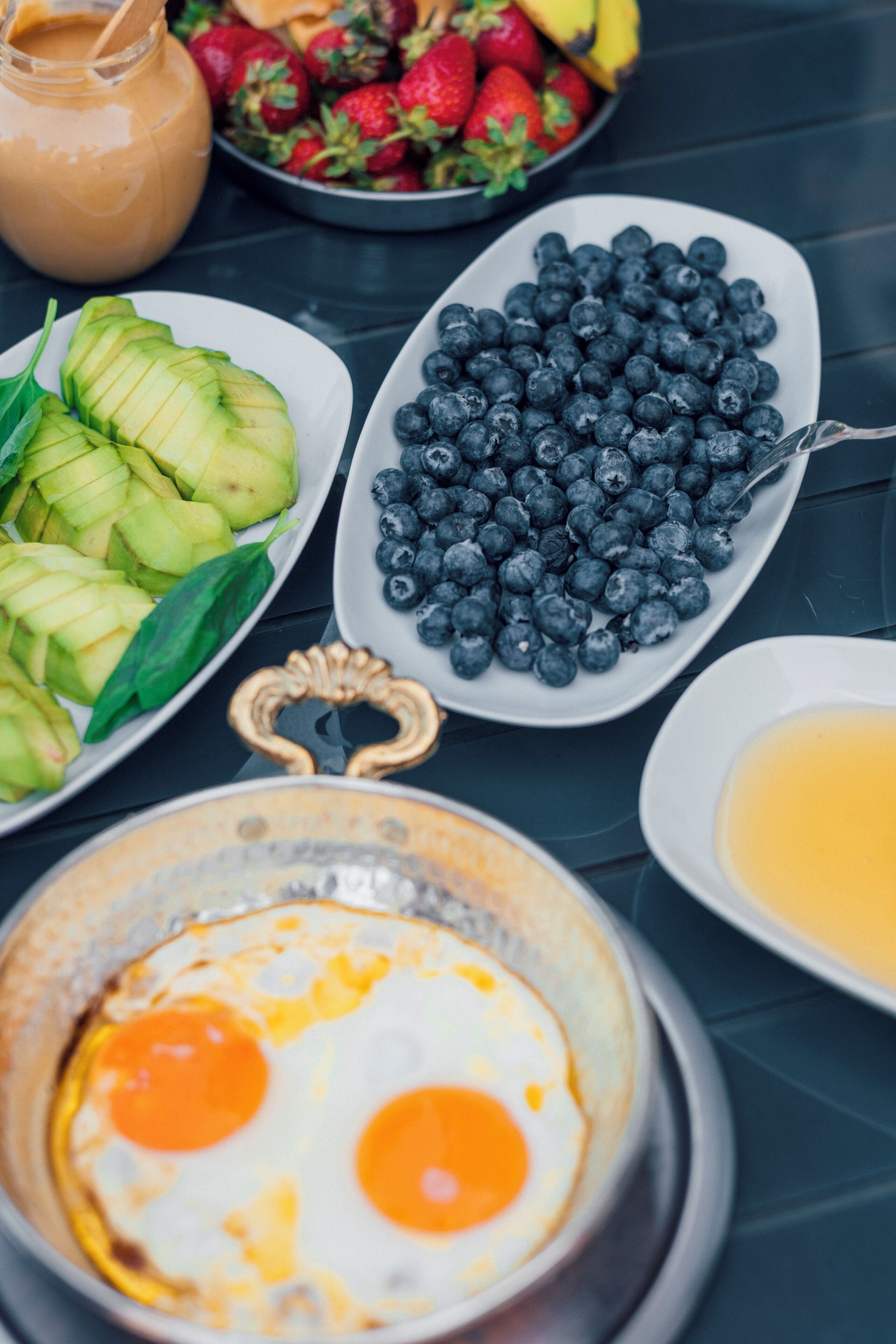 Une table dressée pour le petit-déjeuner avec des tranches d’avocat, des myrtilles, un bol de fraises, un pichet contenant un liquide brun et une assiette avec deux œufs au plat.