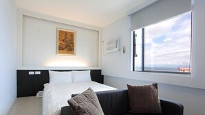 Habitación de hotel con cama, sofá negro, ventana con vistas y un cuadro en la pared.