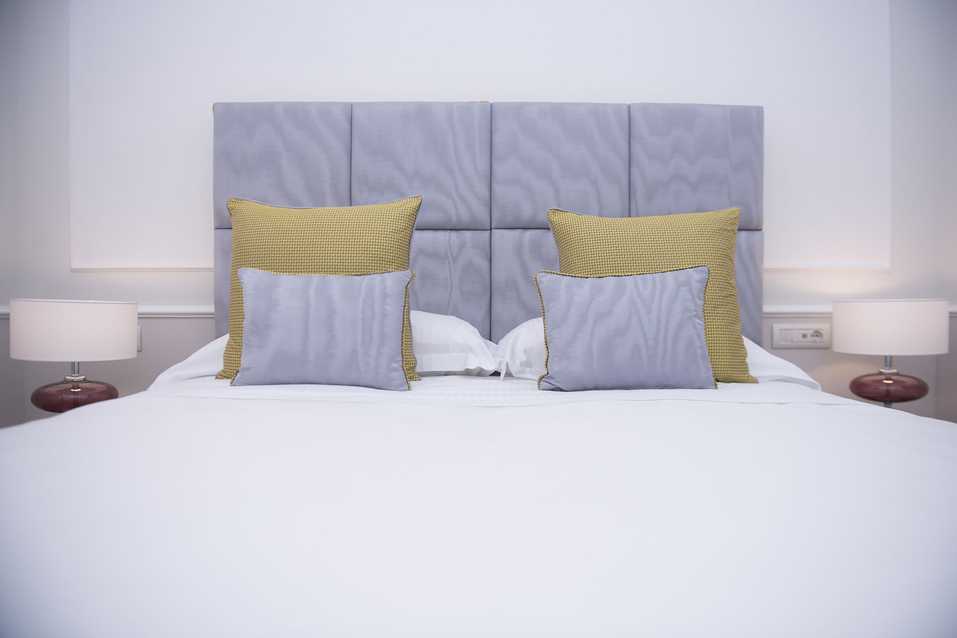 A neatly made bed with a grey upholstered headboard, two pillows with a yellow and grey pattern, and two bedside lamps on either side.