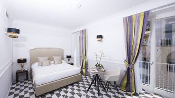 A hotel room with a bed, bedside tables, a small round table with a chair, a potted orchid, and a large window with curtains.