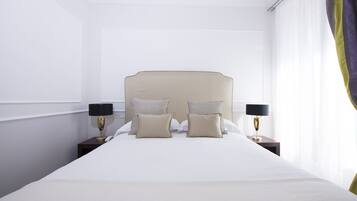 Una cama bien hecha con cabecera, dos mesitas de noche con lámparas y una ventana con cortinas traslúcidas.