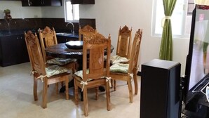 Un coin repas avec une table et des chaises en bois, une cuisine à l’arrière-plan et un téléviseur posé sur un meuble.