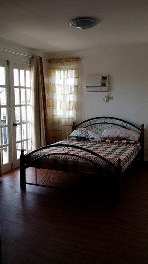 A bedroom with a bed, a window with curtains, an air conditioning unit, and a door.