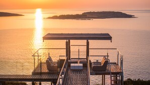 A wooden deck with a sofa and a table, overlooking a calm sea and a distant island at sunset. - Villa Olea Prime (Chania)