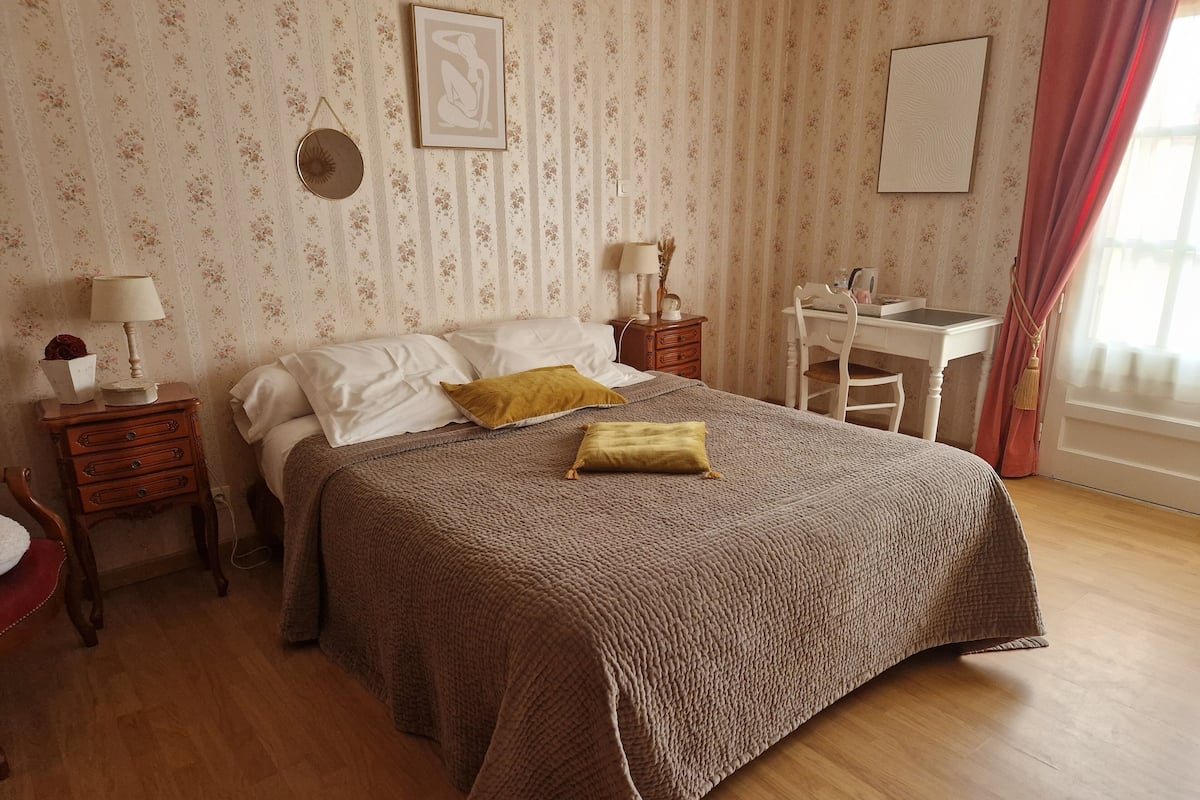 Een slaapkamer met een groot bed, een nachtkastje met een lamp, een kapstok, een bureau en een spiegel.