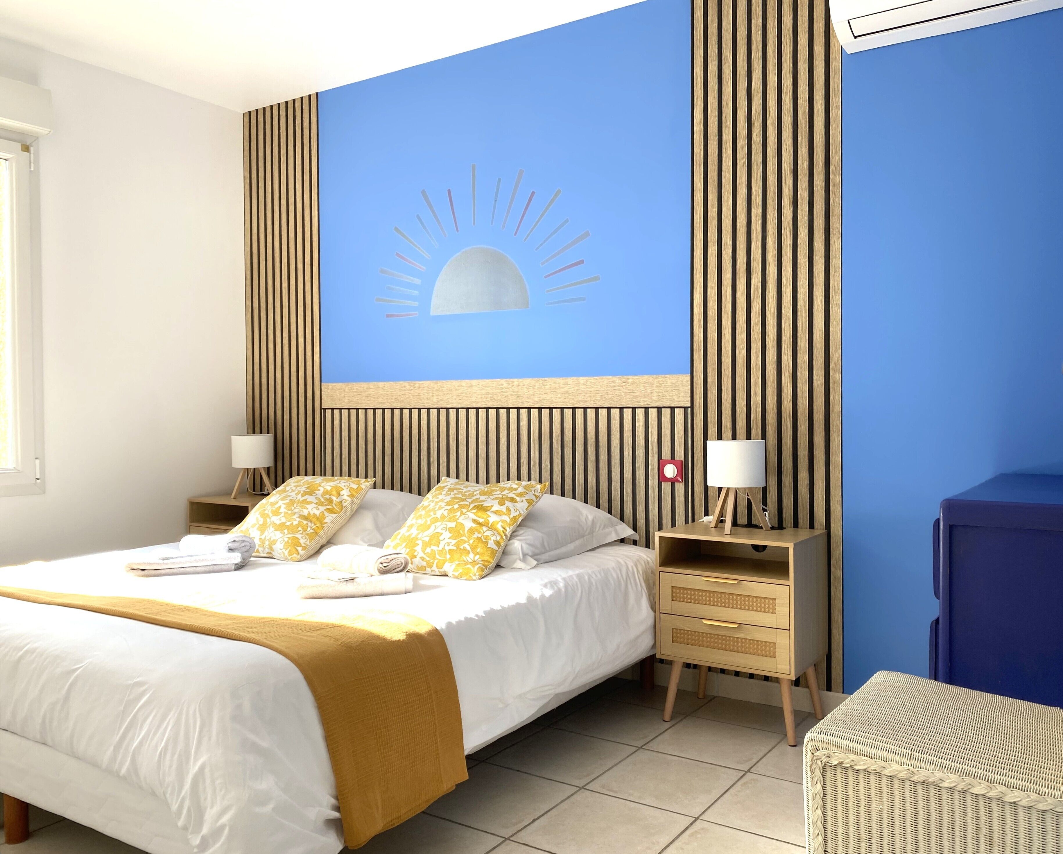 A hotel room with a large bed, two bedside tables, a wall-mounted artwork, and a wicker chair.
