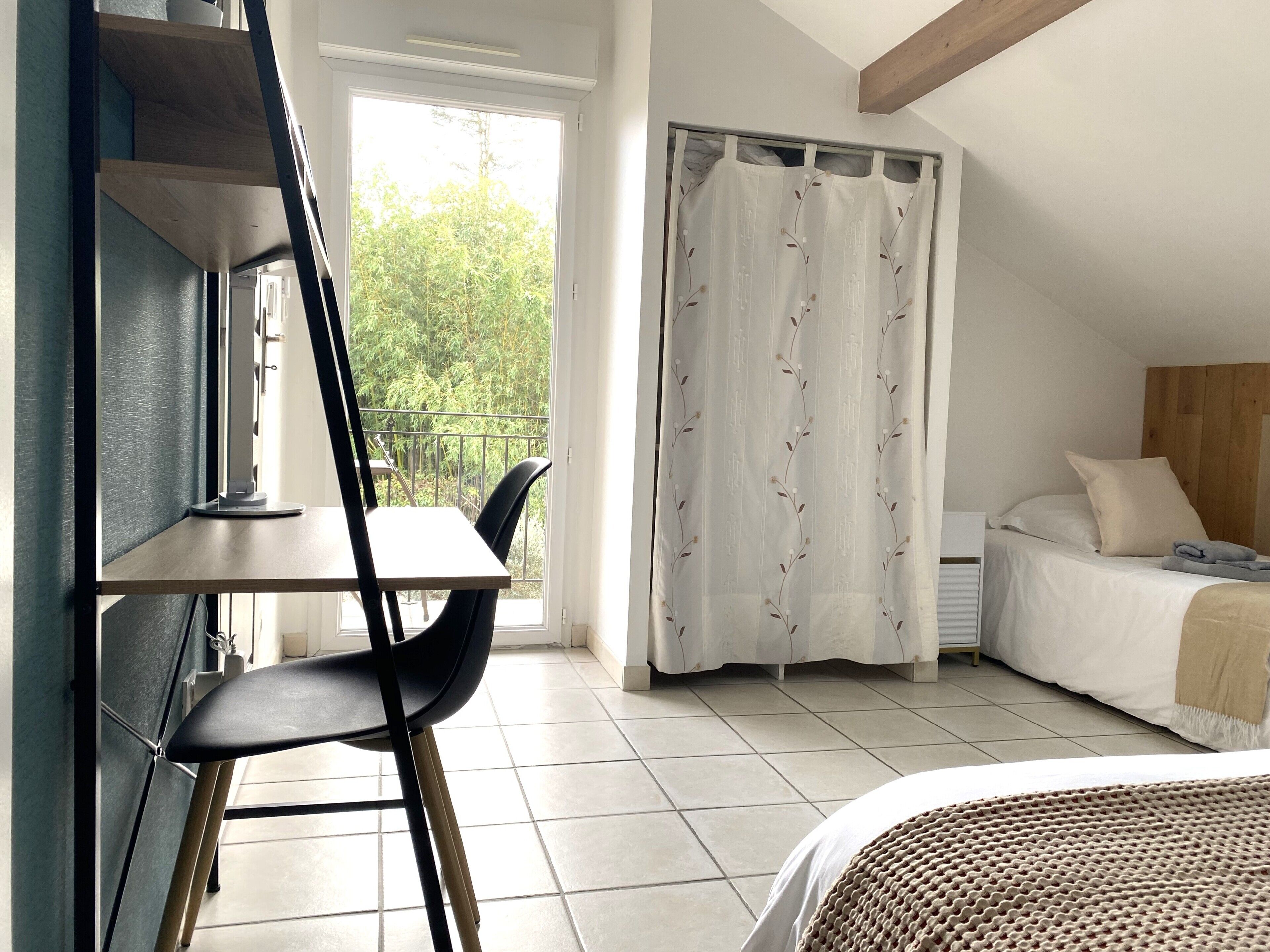 A bedroom with a bed, a desk with a chair, a window with a view of greenery, and a curtain with a floral pattern.