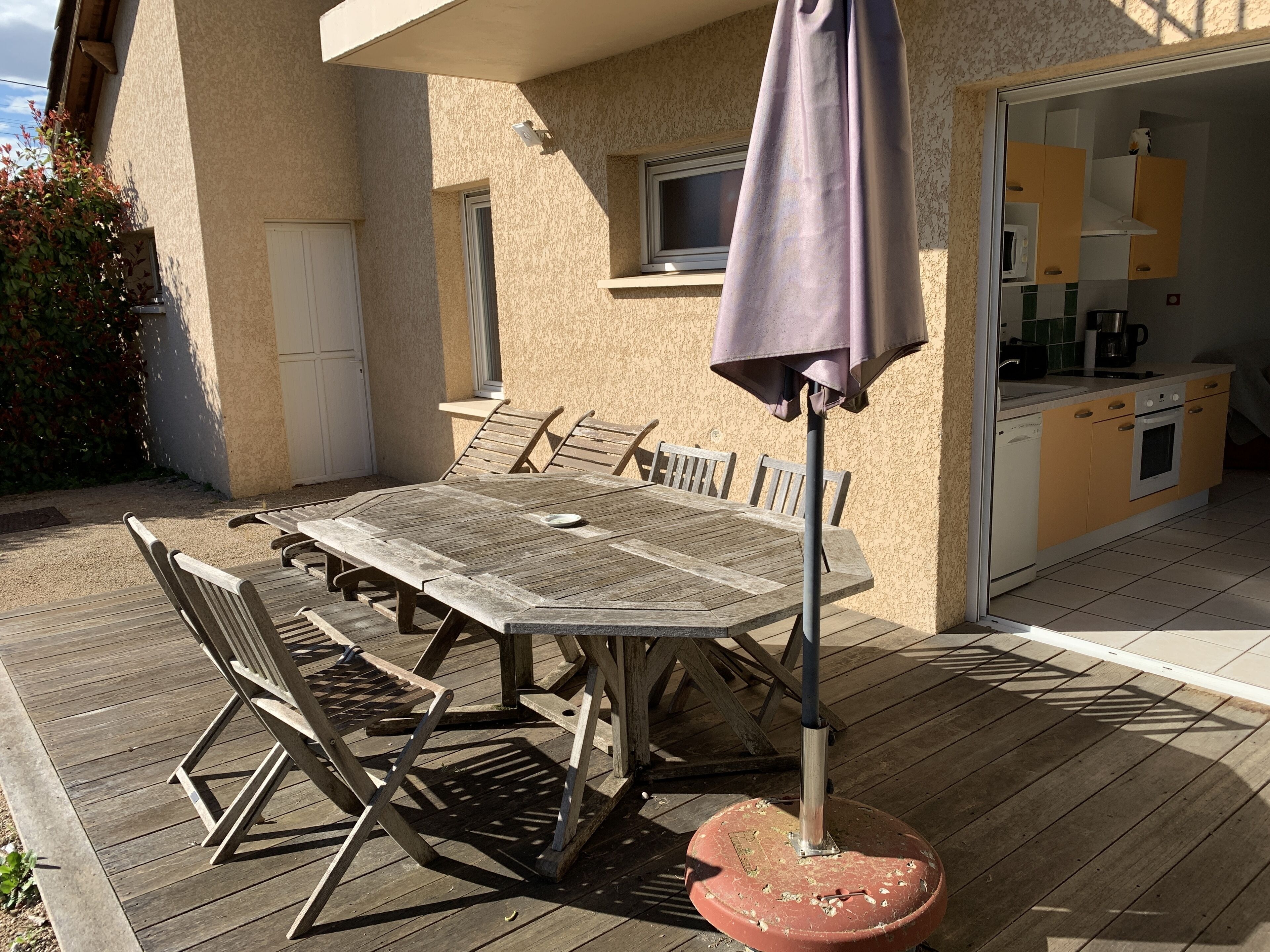 An outdoor patio with a wooden table and chairs, a purple umbrella, and a kitchen area visible through an open door.