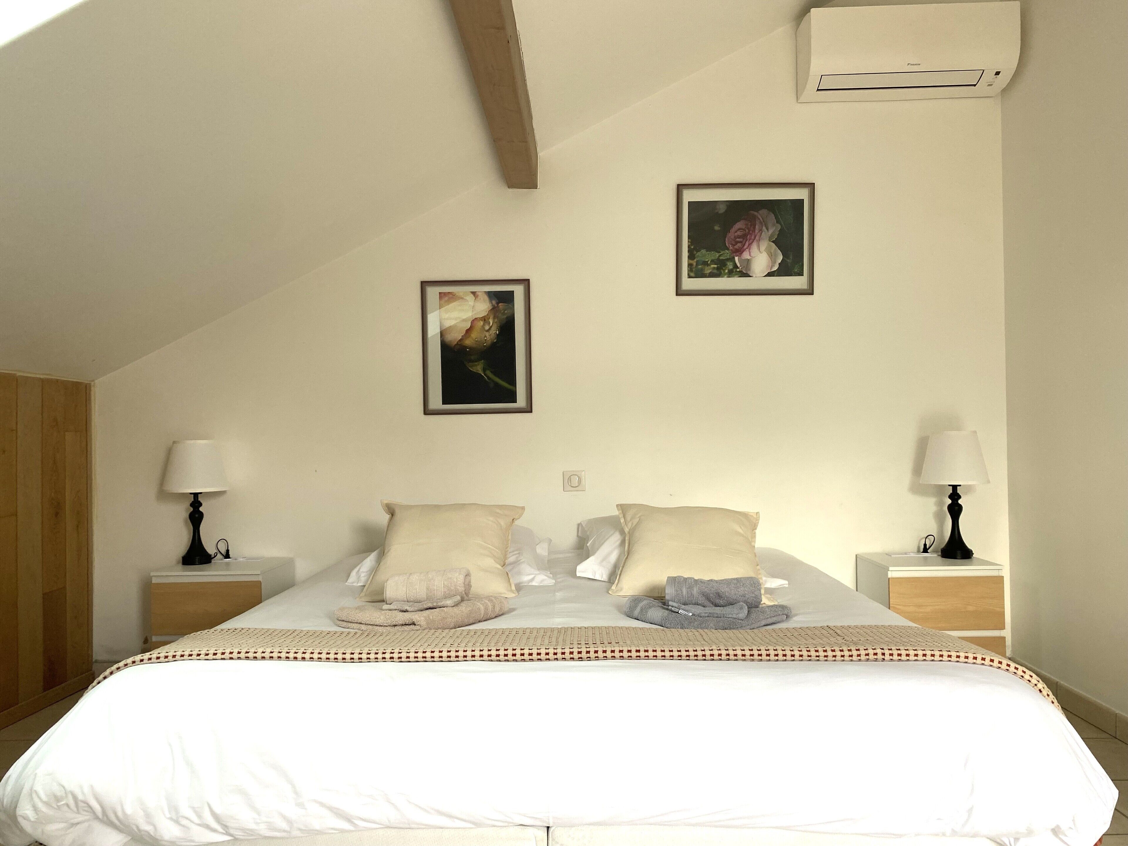 A bedroom with a large bed, two bedside tables with lamps, a framed picture on the wall, and an air conditioning unit.
