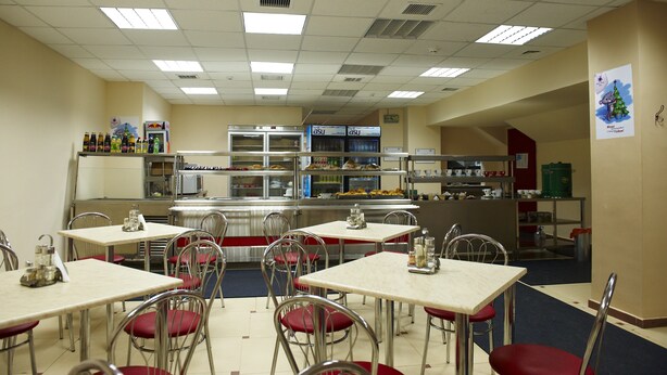 A cafeteria with tables and chairs, a kitchen area, and a clock on the wall.