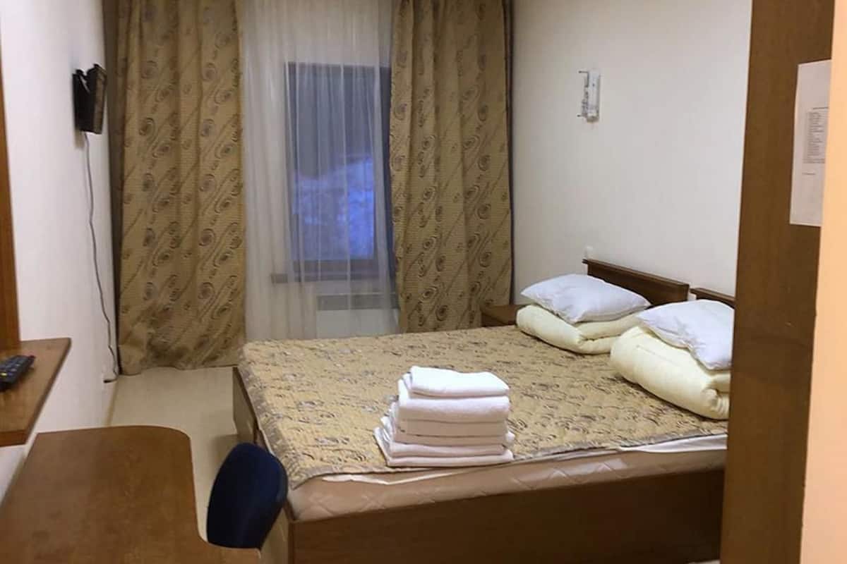 A small, neatly arranged bedroom with a bed, pillows, a bedside table, and a window with curtains.