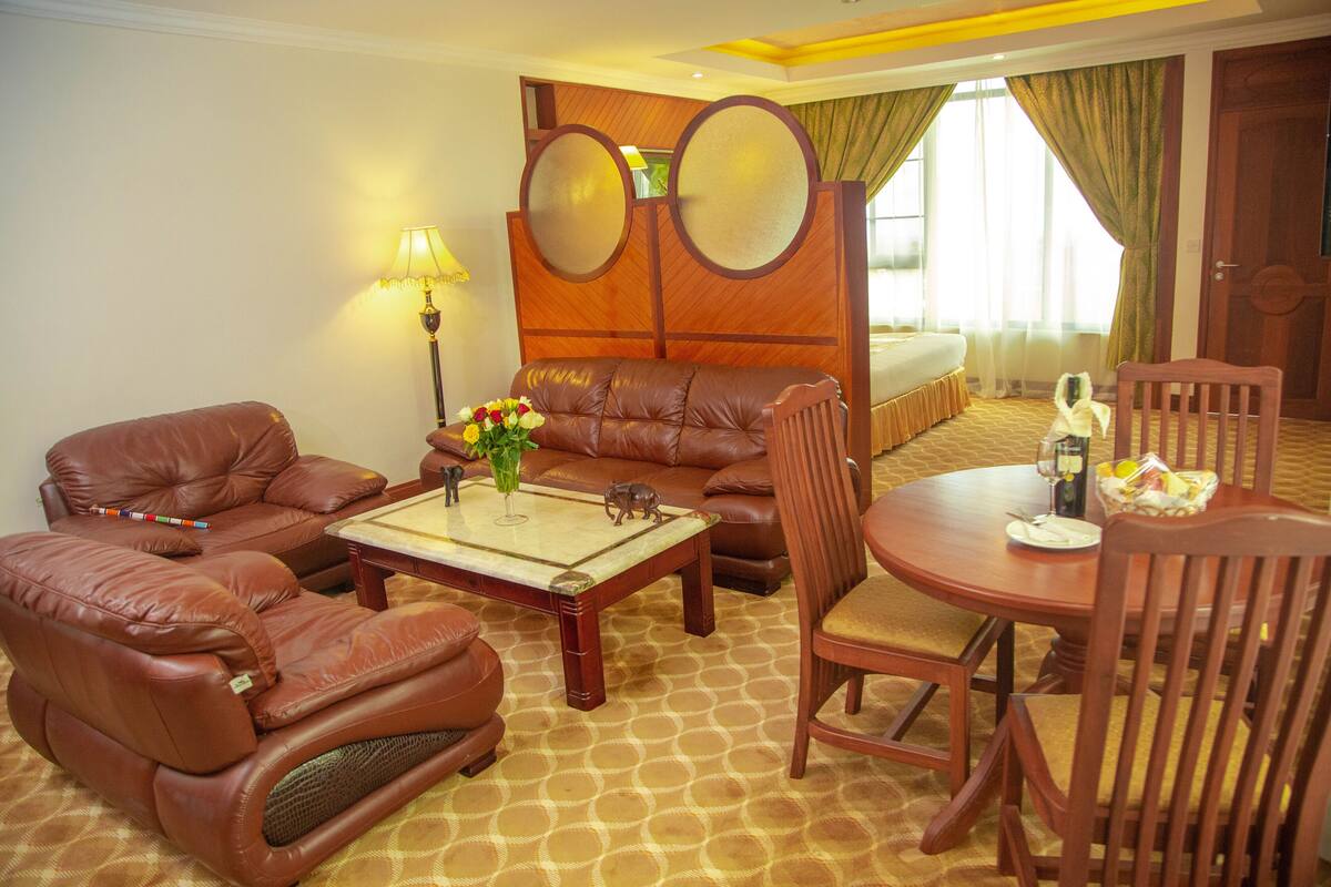 A hotel room with a brown leather sofa, a wooden coffee table, a dining table with chairs, and a bed with a wooden headboard.