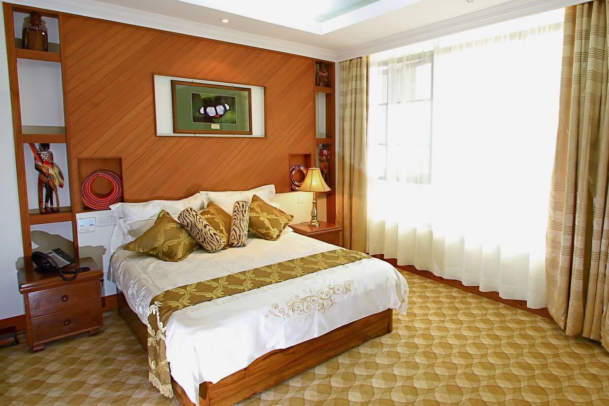 A hotel room with a large bed, a wooden headboard, a framed picture on the wall, a bedside table with a lamp, and a patterned carpet.