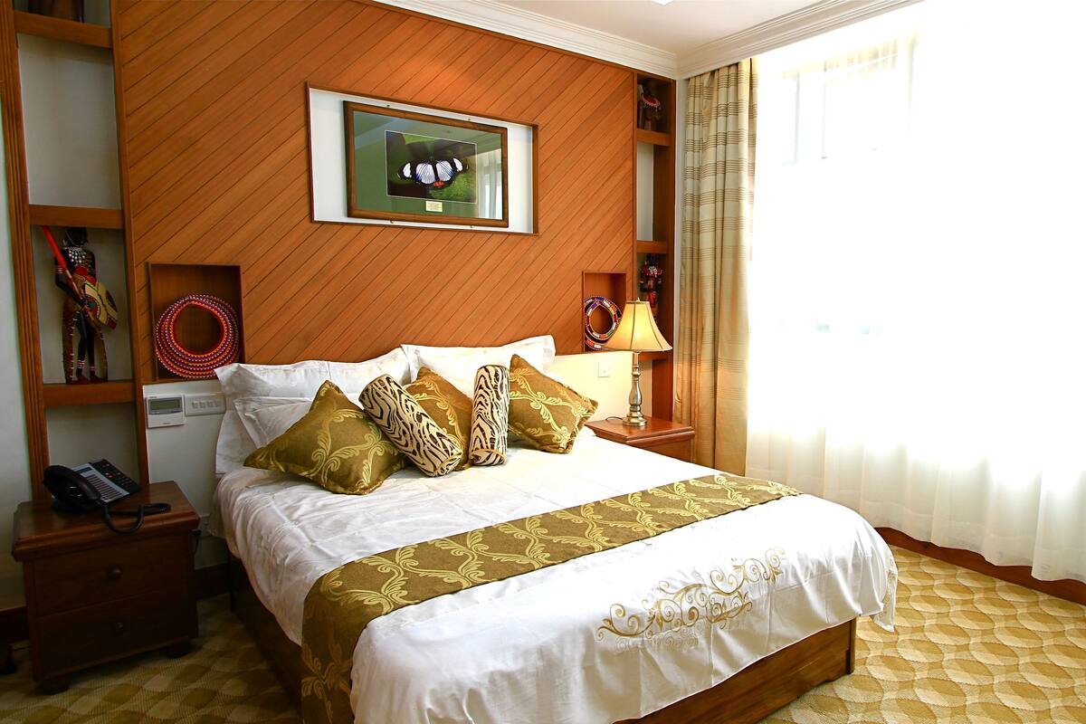 A hotel room with a bed, a nightstand with a phone, a lamp, a framed picture on the wall, and a window with curtains.