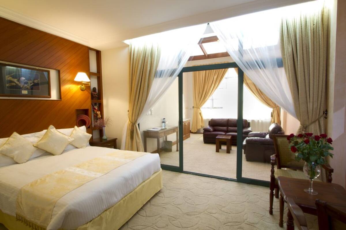 A hotel room with a bed, a sitting area with a sofa and armchairs, a dining table, and a balcony with curtains.