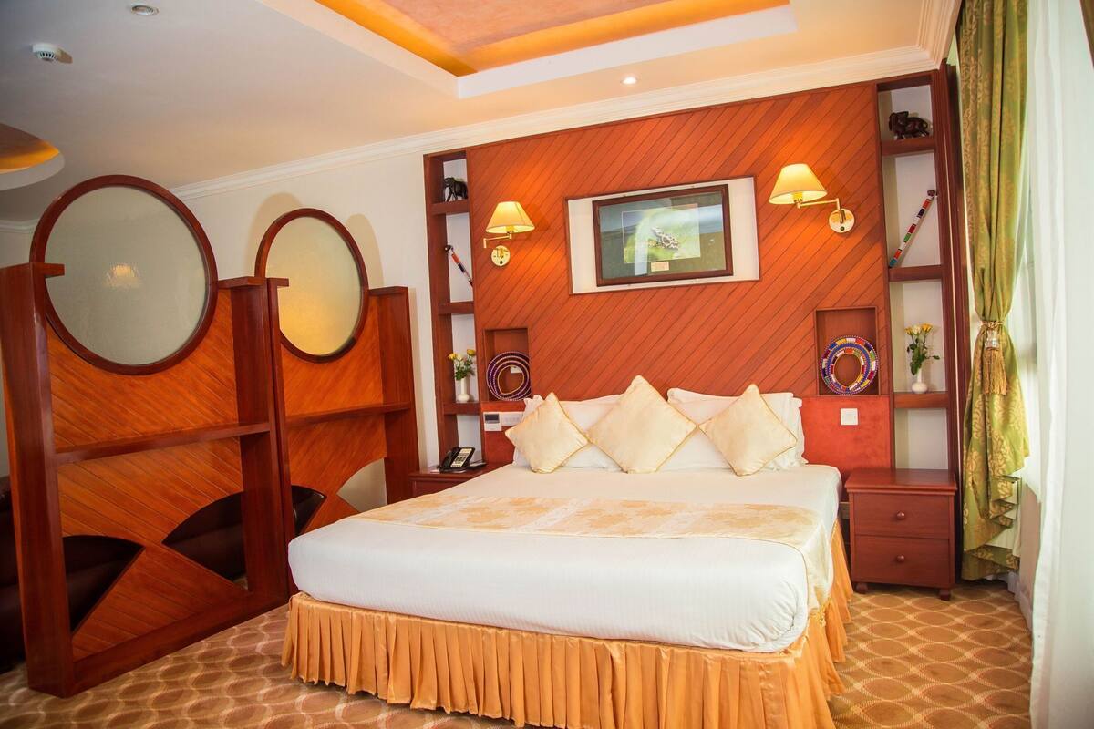 A hotel room with a large bed, a wooden headboard, a mirror, a bedside table, a nightstand, and a wall-mounted TV.