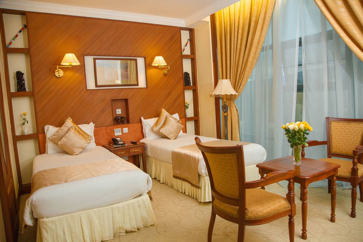 A hotel room with two beds, a desk with a chair, a lamp, and a vase with flowers.
